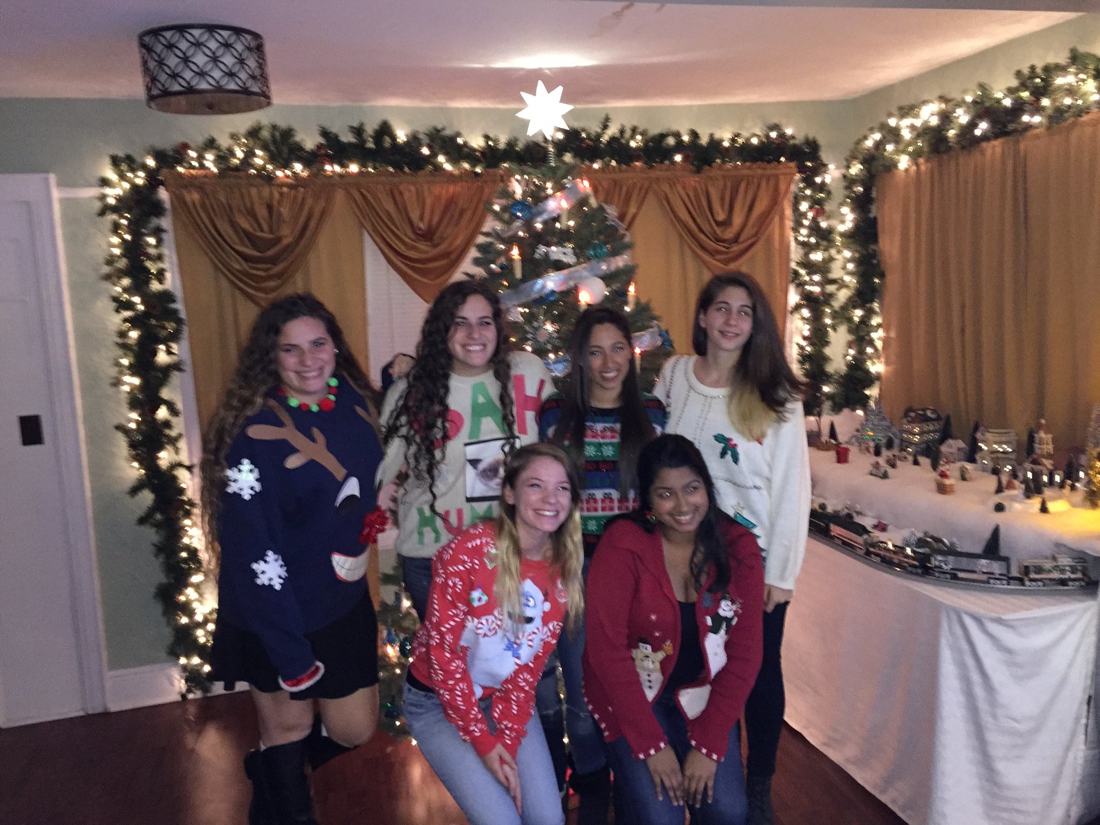 Six friends in festive holiday sweaters beside a decorated Christmas tree